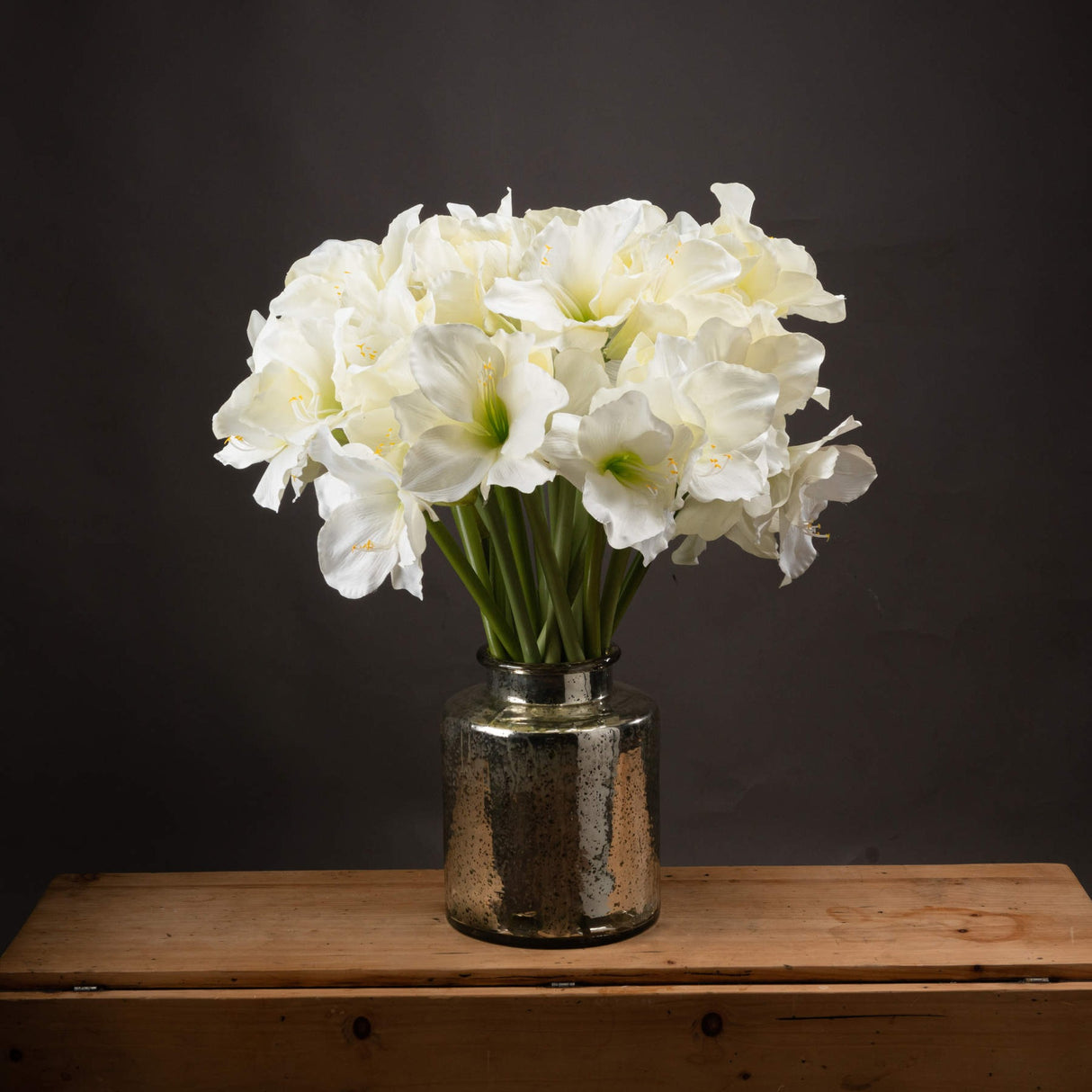 Hill Interiors Classic White Amaryllis faux flower stems arranged in a mercury glass vase on a wooden surface against a dark grey background.