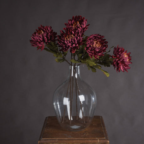 Hill Interiors Autumn Spider Chrysanthemum faux stems with purple shaggy blooms and green foliage arranged in a clear glass vase on a wooden stand.