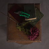Hill Interiors Autumn Spider Chrysanthemum faux stem with purple petals and green foliage laid on a wooden table beside vintage books and green secateurs.