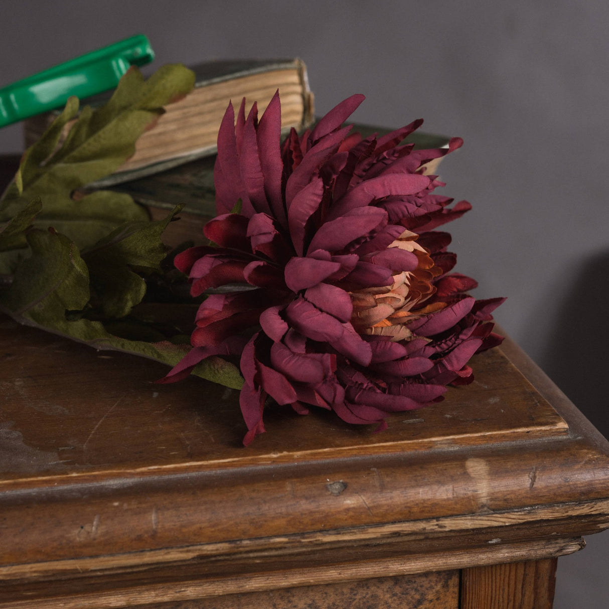 Hill Interiors Autumn Spider Chrysanthemum faux stem with purple layered petals and green foliage, resting on a wooden console beside antique books.