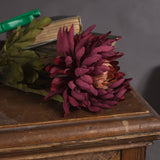 Hill Interiors Autumn Spider Chrysanthemum faux stem with purple layered petals and green foliage, resting on a wooden console beside antique books.