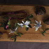 Hill Interiors Passion Flower Spray with white blooms and green foliage laid on a wooden table beside a burgundy ribbon and pine cones.