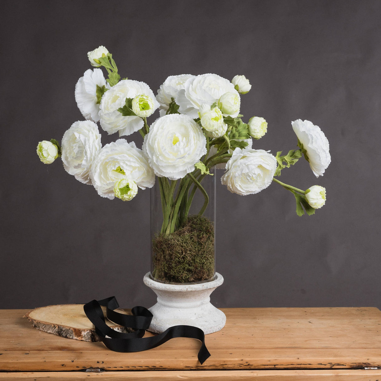 Hill Interiors White Ranunculus Spray arranged with buds and open blooms in a glass vase with moss on a stone plinth against a grey backdrop.