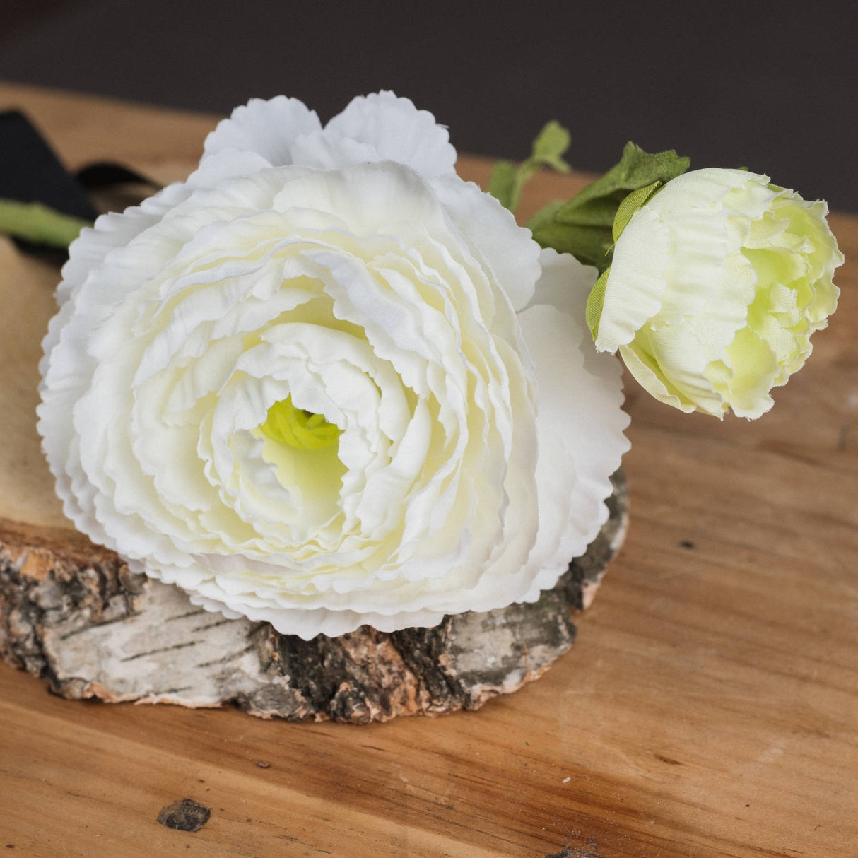 Hill Interiors White Ranunculus Spray with a full open bloom and a smaller pale green bud, displayed on a birch wood slice atop a wooden surface.