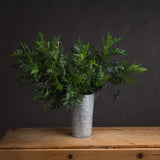 Hill Interiors Pistach Bunch faux foliage stems with rounded green leaves arranged in a tall galvanised metal florist bucket on a wooden surface.