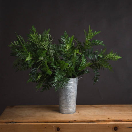 Hill Interiors Pistach Bunch faux foliage stems with rounded green leaves arranged in a tall galvanised metal florist bucket on a wooden surface.
