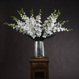 White Gladioli faux flower stems by Hill Interiors arranged in a hammered silver vase on a wooden side table against a dark background.