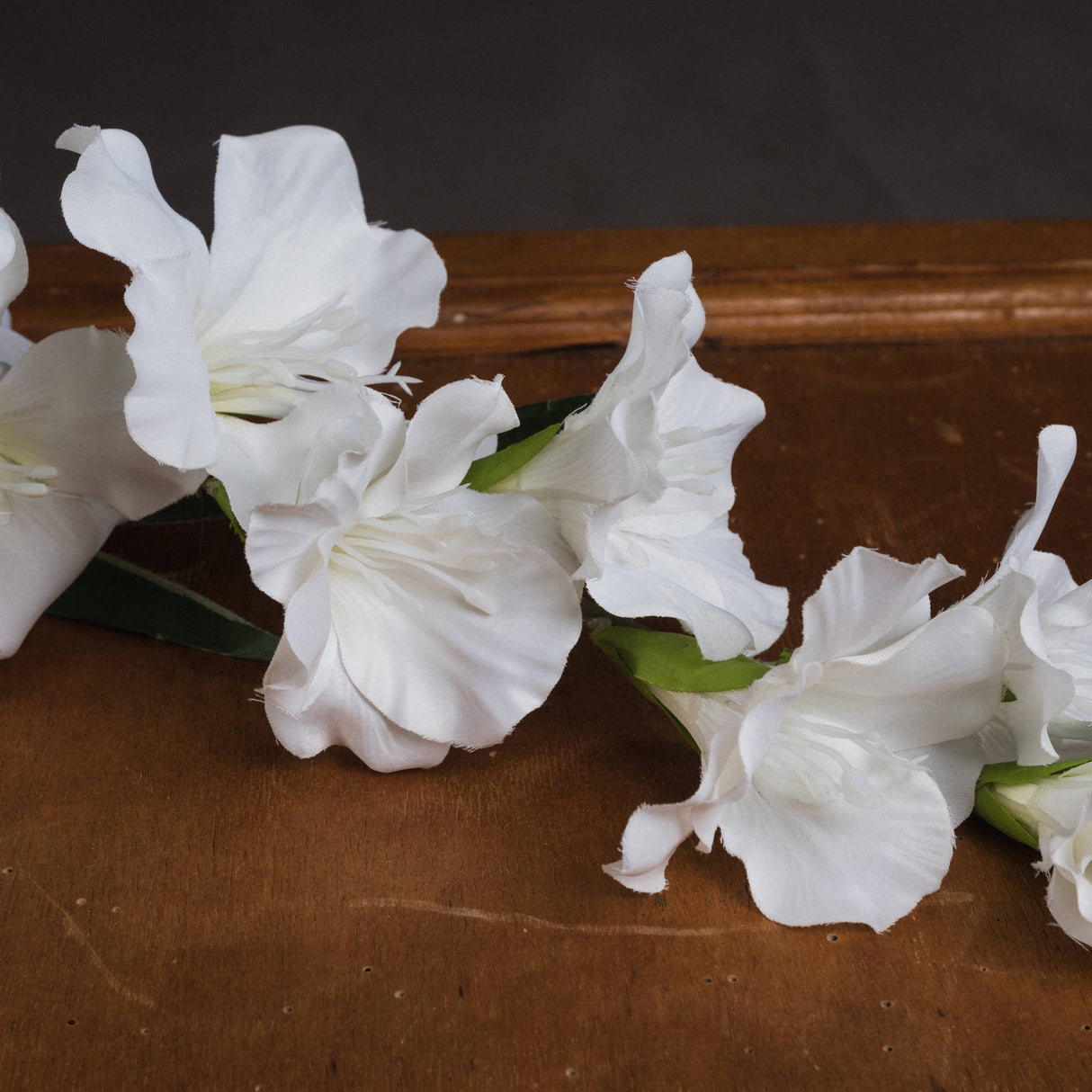 Hill Interiors White Gladioli faux flower stem with ruffled white blooms and green leaves, displayed on a wooden surface against a dark background.