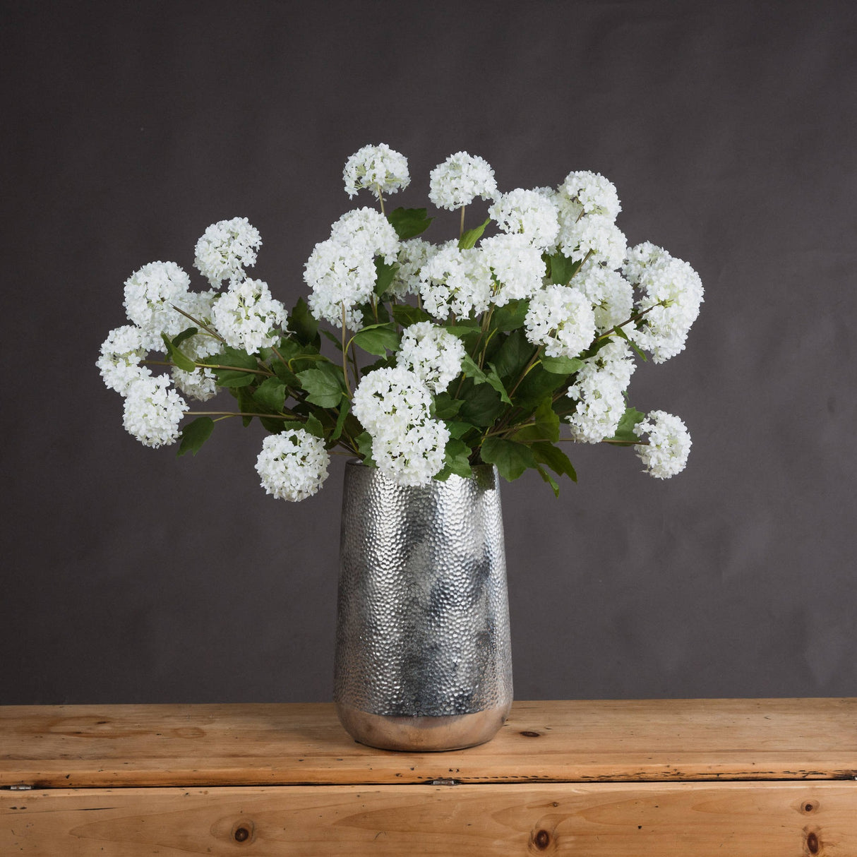 Hill Interiors Viburnum Opulus faux flower stems with white spherical blooms and green leaves, arranged in a hammered silver vase on a wooden surface.