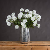 Hill Interiors Viburnum Opulus faux flower stems with white spherical blooms and green leaves, arranged in a hammered silver vase on a wooden surface.
