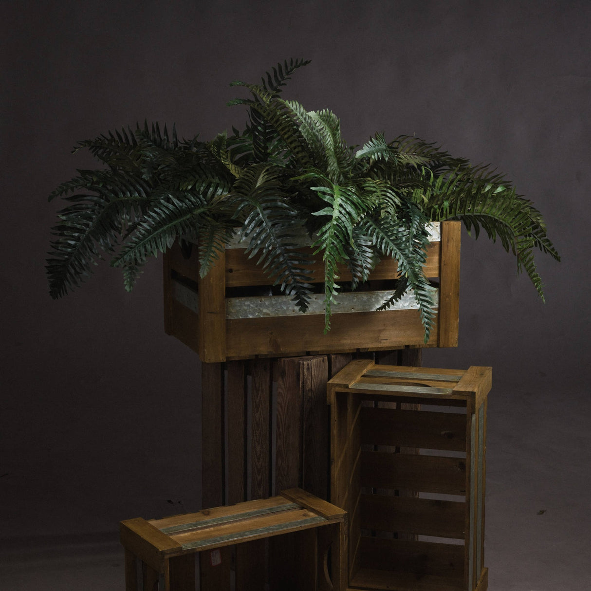 Hill Interiors Fern Bunch faux foliage with arching green fronds displayed in a wooden crate atop stacked rustic crates against a dark grey backdrop.