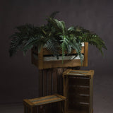 Hill Interiors Fern Bunch faux foliage with arching green fronds displayed in a wooden crate atop stacked rustic crates against a dark grey backdrop.
