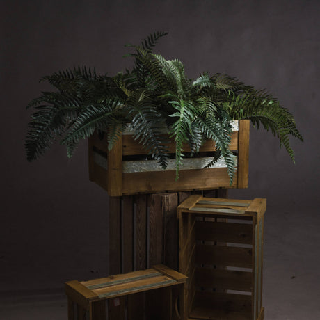 Hill Interiors Fern Bunch faux foliage with arching green fronds displayed in a wooden crate atop stacked rustic crates against a dark grey backdrop.