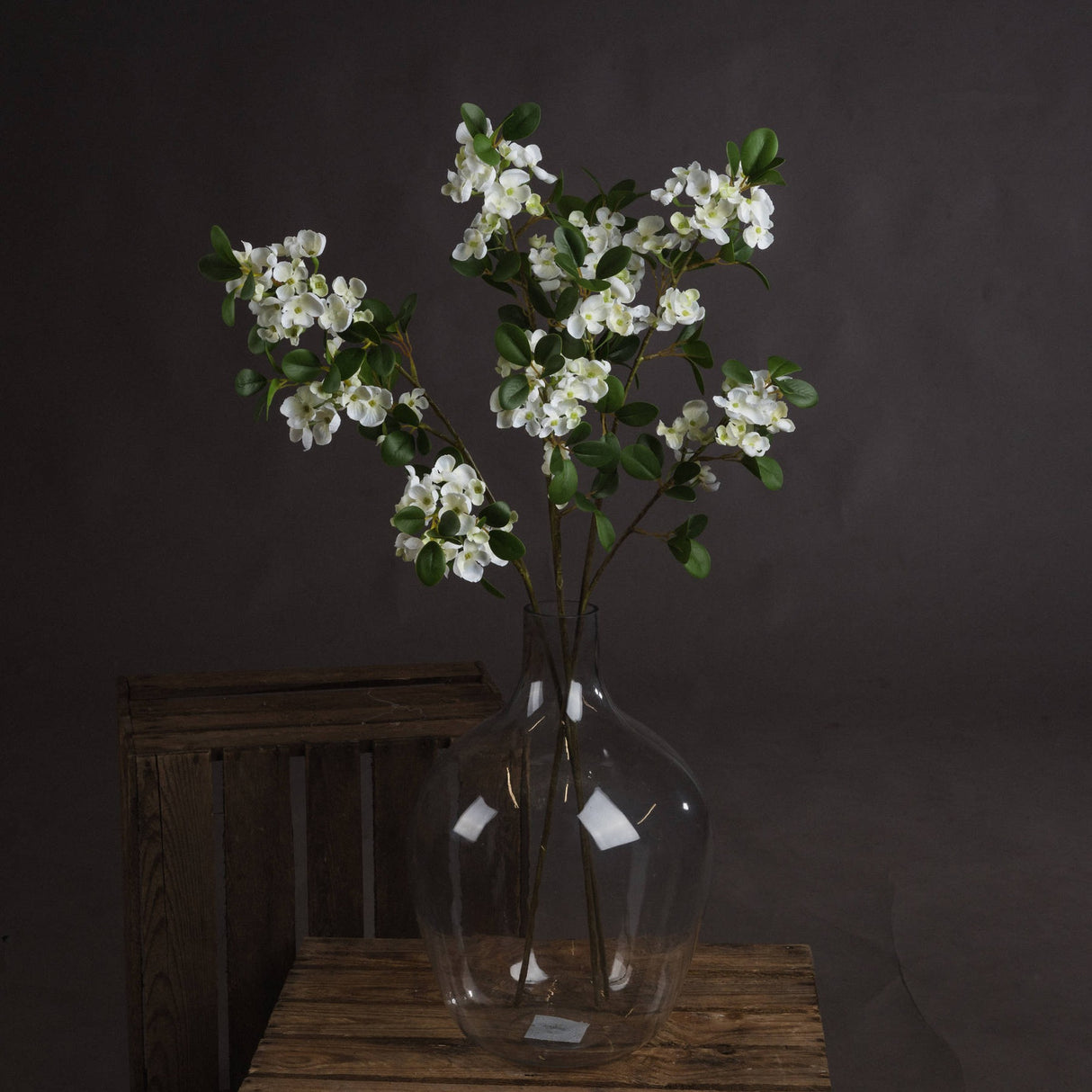 Hill Interiors White Spring Mock Orange Flower stems with cupped white blossoms and green leaves arranged in a clear glass vase on a wooden crate.