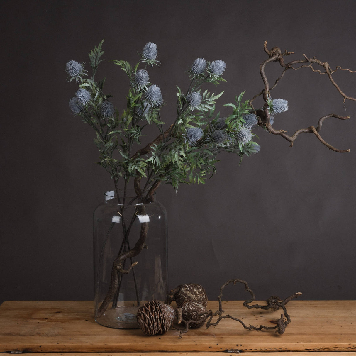 Hill Interiors Eryngium Thistle faux stems with grey cone heads and green bracts in a glass jar with twisted branches and pine cones.