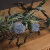 Hill Interiors Eryngium Thistle faux stem with grey cone-shaped thistle heads and jagged green bracts, displayed on a wooden surface with twisted branches.
