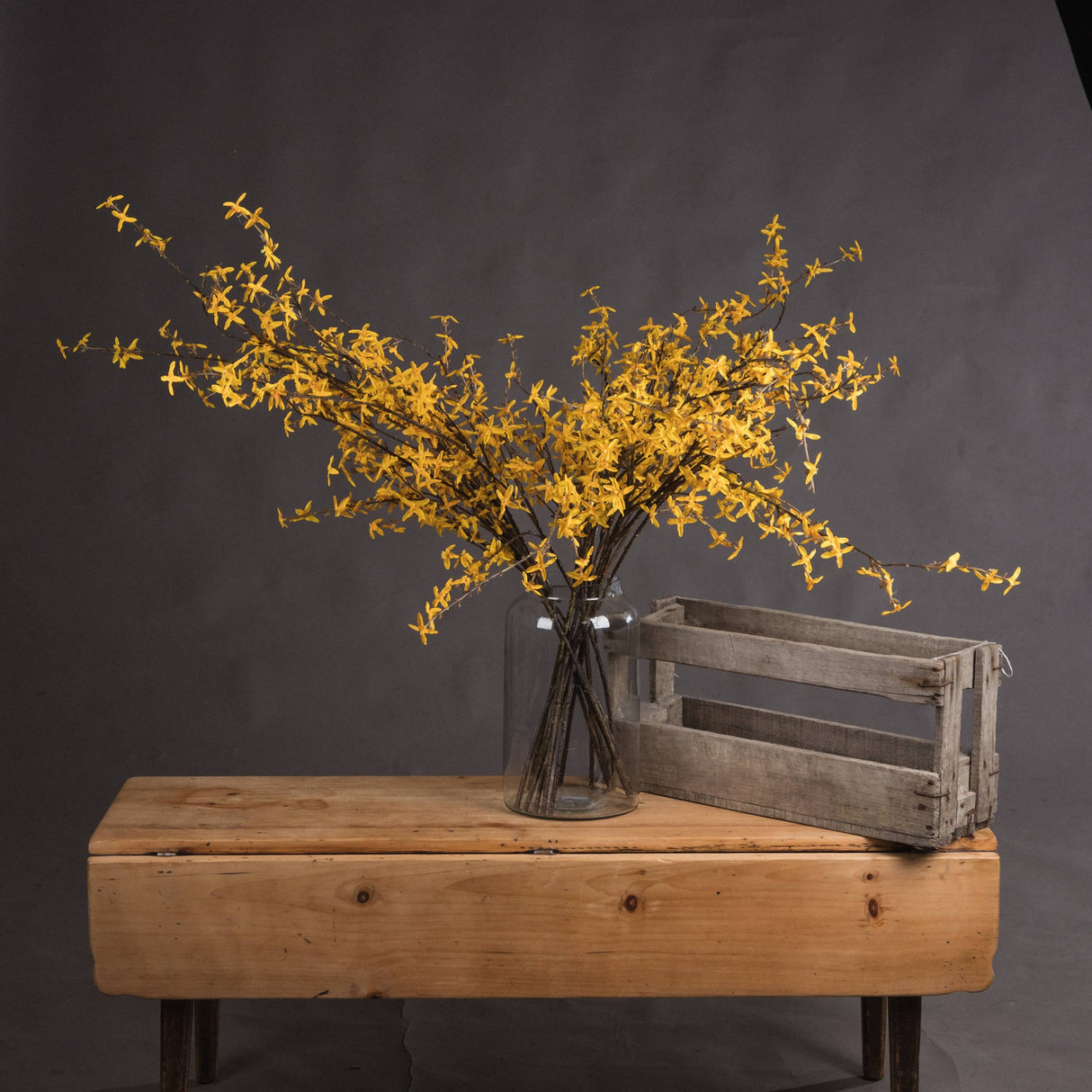 Hill Interiors Forsythia faux flower stems with yellow blooms on woody branches, arranged in a clear glass jar vase beside a wooden crate on a pine bench.
