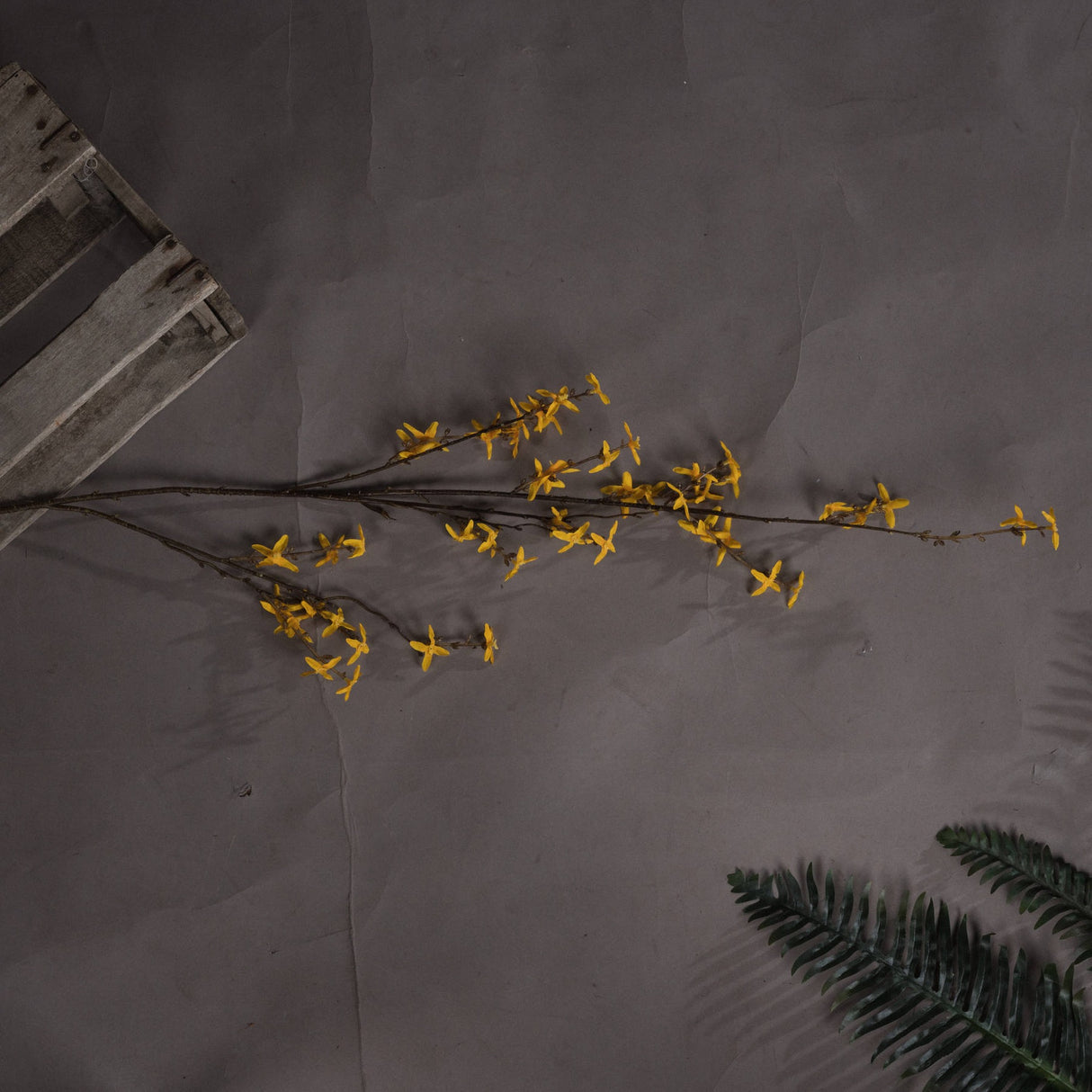 Hill Interiors Forsythia faux flower stem with clusters of yellow four-petalled blooms on woody branches, laid on grey with a wooden crate and fern.