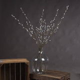 Hill Interiors Pussy Willow Branch faux foliage stems with downy pale buds arranged in a clear glass vase on a wooden crate against a grey backdrop.