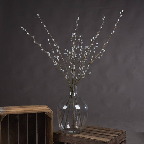 Hill Interiors Pussy Willow Branch faux foliage stems with downy pale buds arranged in a clear glass vase on a wooden crate against a grey backdrop.