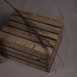 Hill Interiors Branch Spray faux foliage stem in warm brown plastic, laid diagonally across stacked wooden slatted crates on a grey backdrop.