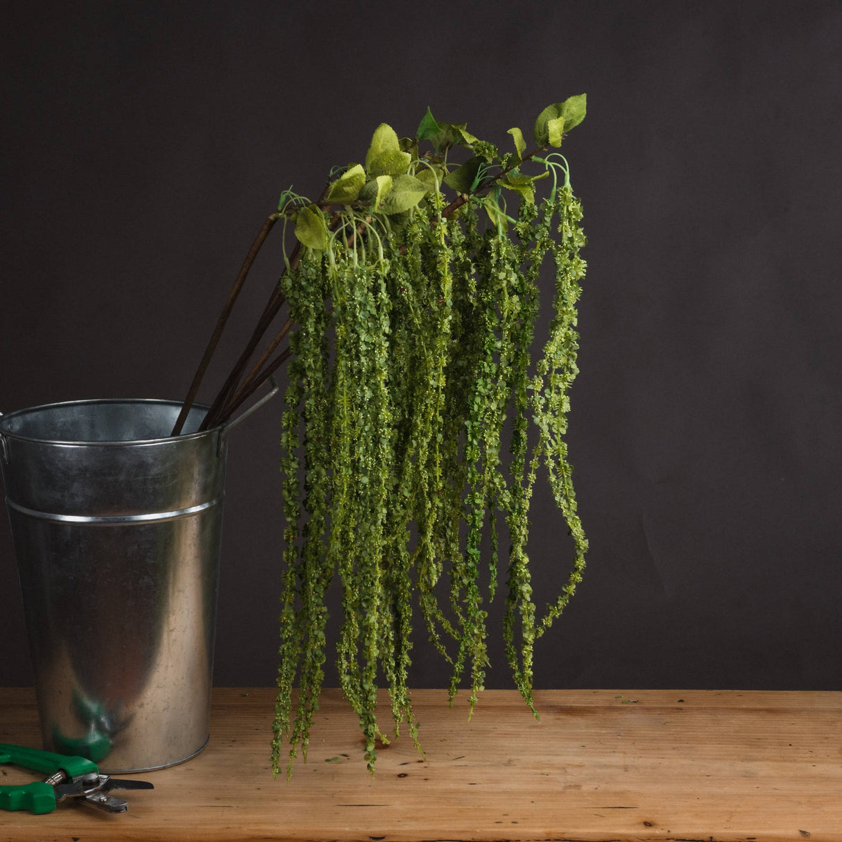 Hill Interiors Green Amaranthus faux stem with cascading beaded plumes displayed in a galvanised metal bucket on a wooden surface against a dark background.