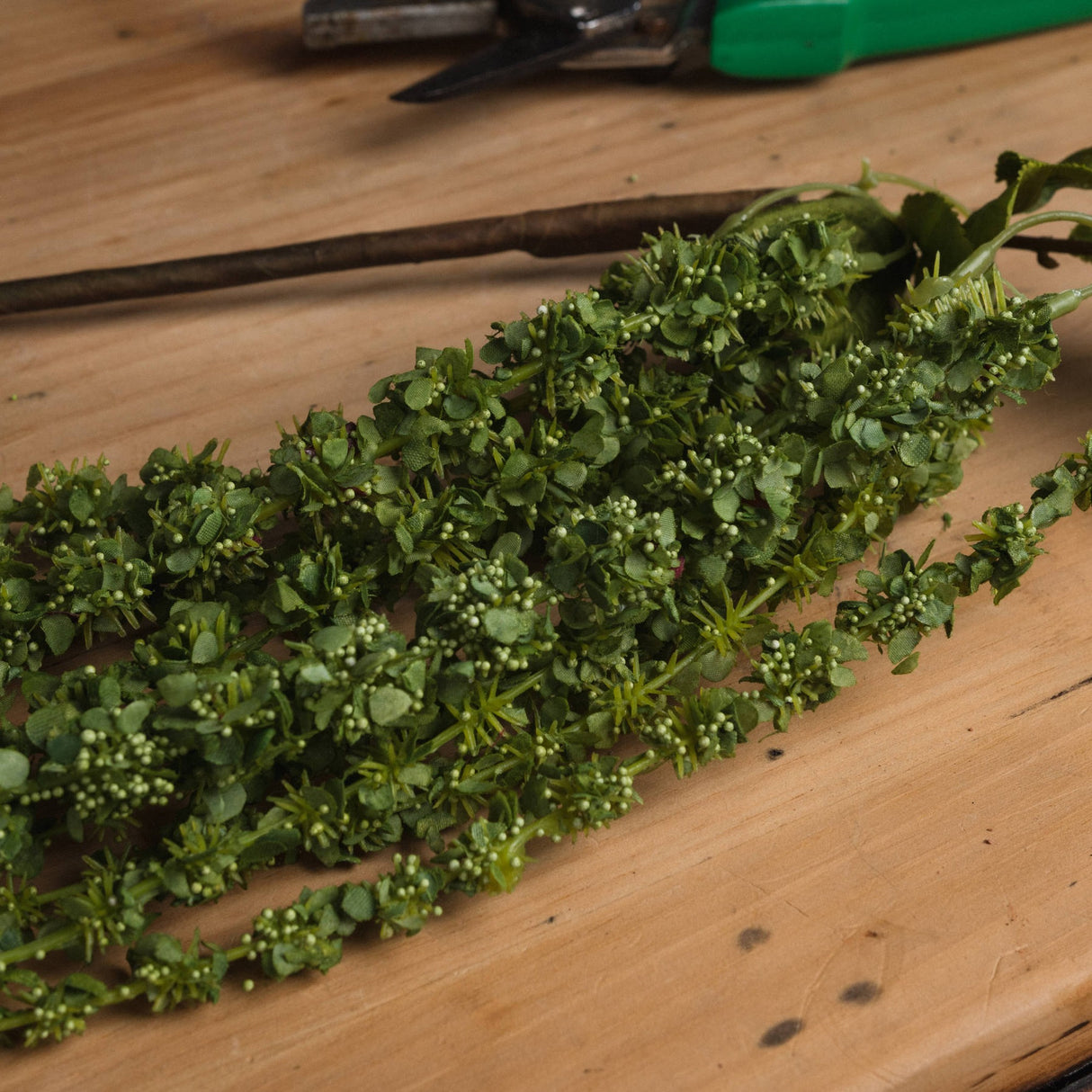 Hill Interiors Green Amaranthus faux flower stem with trailing beaded plumes and slim leaves, resting on a wooden surface near garden snips.