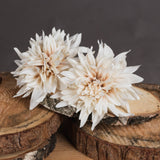 Hill Interiors Café Au Lait faux dahlia stems with layered cream and blush petals, displayed on stacked wooden log slices against a dark grey background.