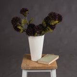Hill Interiors Chocolate Chrysanthemum faux stems with deep brown layered blooms and green leaves, arranged in a distressed white metal vase on a wooden stool.
