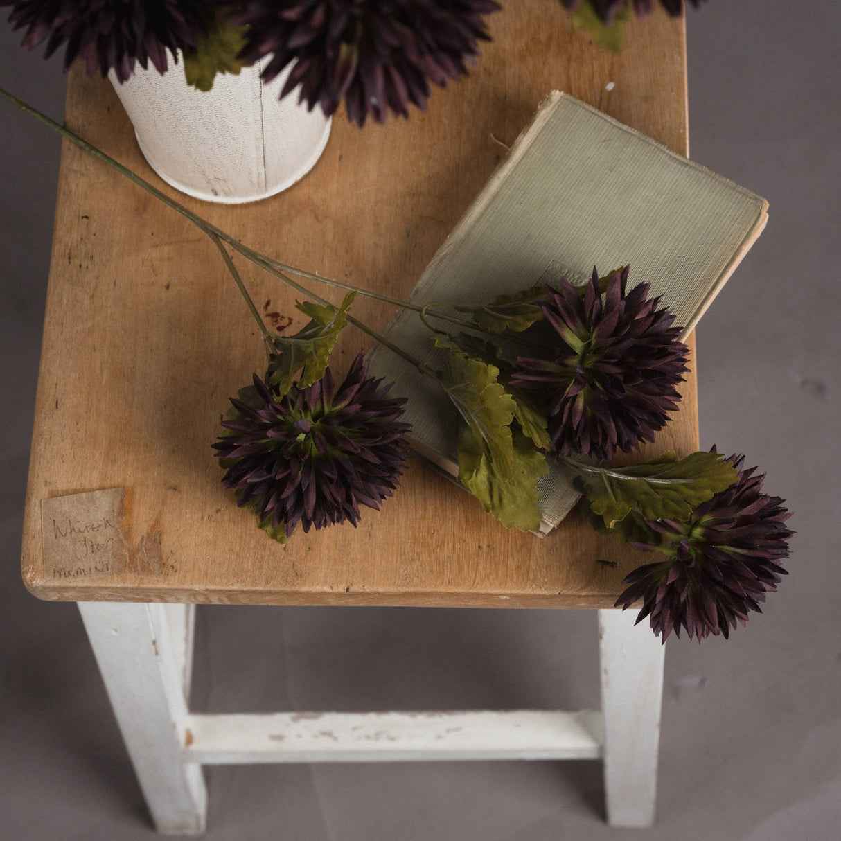 Hill Interiors Chocolate Chrysanthemum faux stems with deep brown layered blooms and green leaves arranged on a wooden stool with a vintage book and vase.