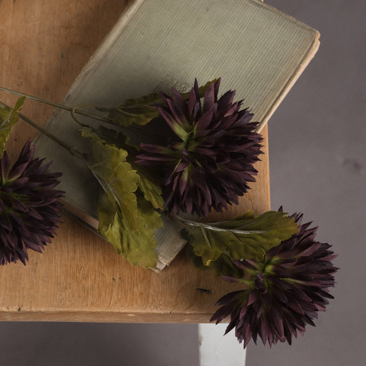 Hill Interiors Chocolate Chrysanthemum faux stems with deep brown layered petals and green leaves resting on a vintage book atop a wooden stool.