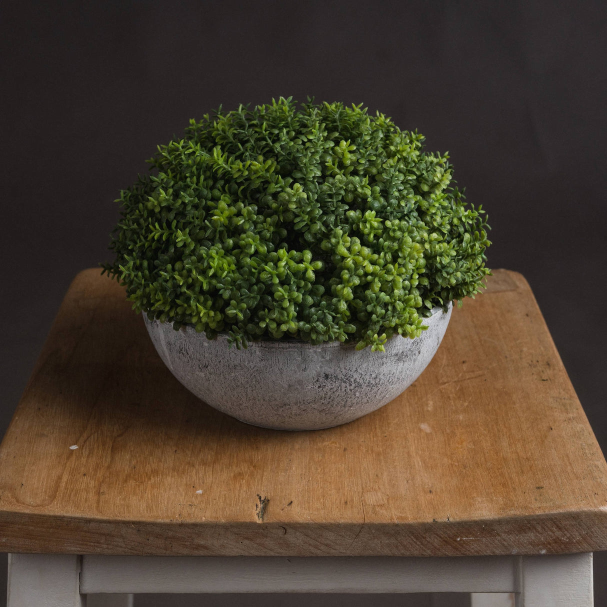 Hill Interiors Small Hebe Globe Pot with a dense green faux foliage dome in a distressed grey stone-effect bowl, displayed on a wooden stool.