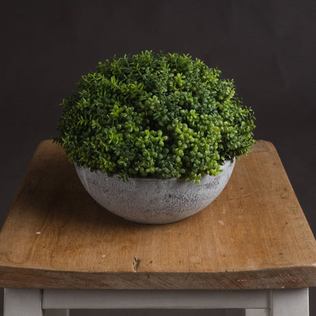 Hill Interiors Small Hebe Globe Pot with a dense green faux foliage dome in a distressed grey stone-effect bowl, displayed on a wooden stool.