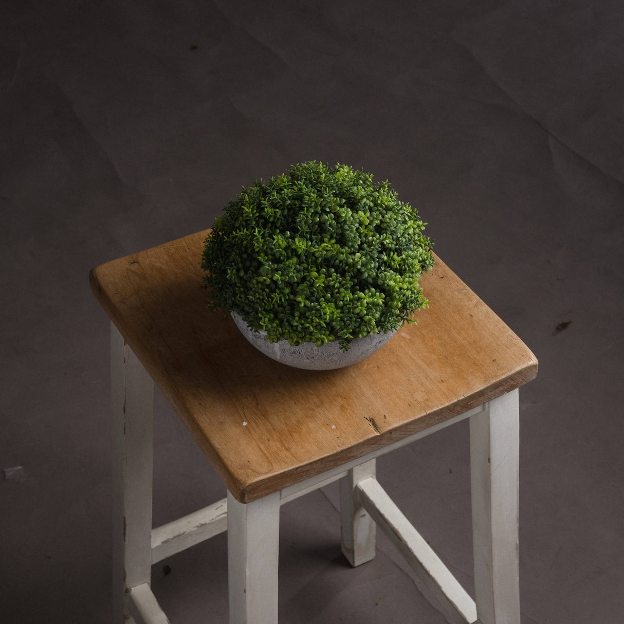 Hill Interiors Small Hebe Globe Pot with dense green faux foliage in a textured grey bowl, displayed on a rustic wooden stool against a dark backdrop.