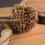 Hill Interiors Pink Allium faux stem with a rounded globe head of blush and rose florets on a green stem, resting on rustic wooden boards.