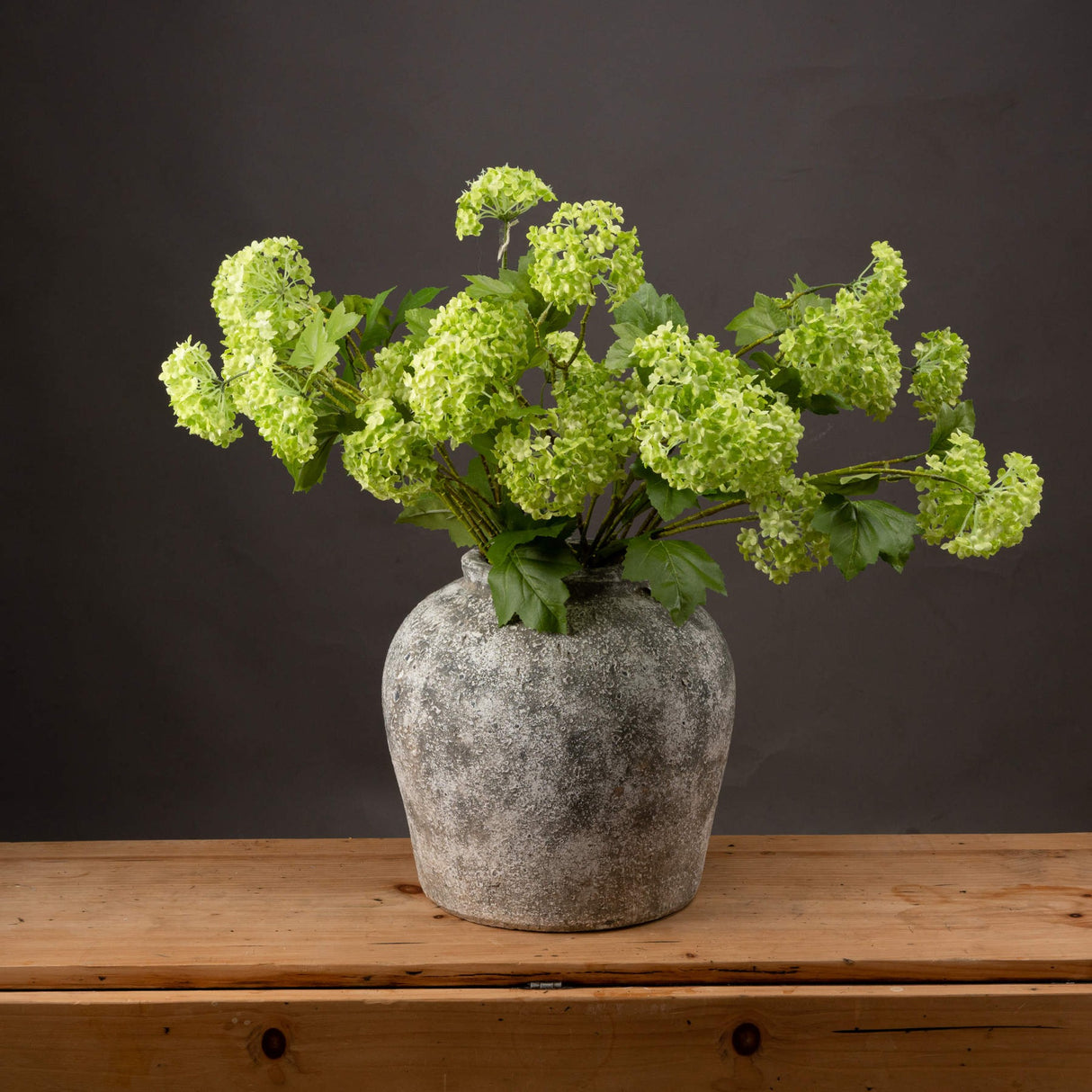 Hill Interiors Green Viburnum Spray arrangement with domed pale green flower clusters and veined leaves in a weathered stone vase on a wooden surface.
