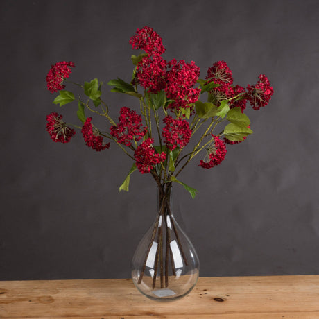 Hill Interiors Red Autumnal Viburnum faux flower stems with clustered red blooms and green leaves arranged in a clear glass vase on a wooden surface.