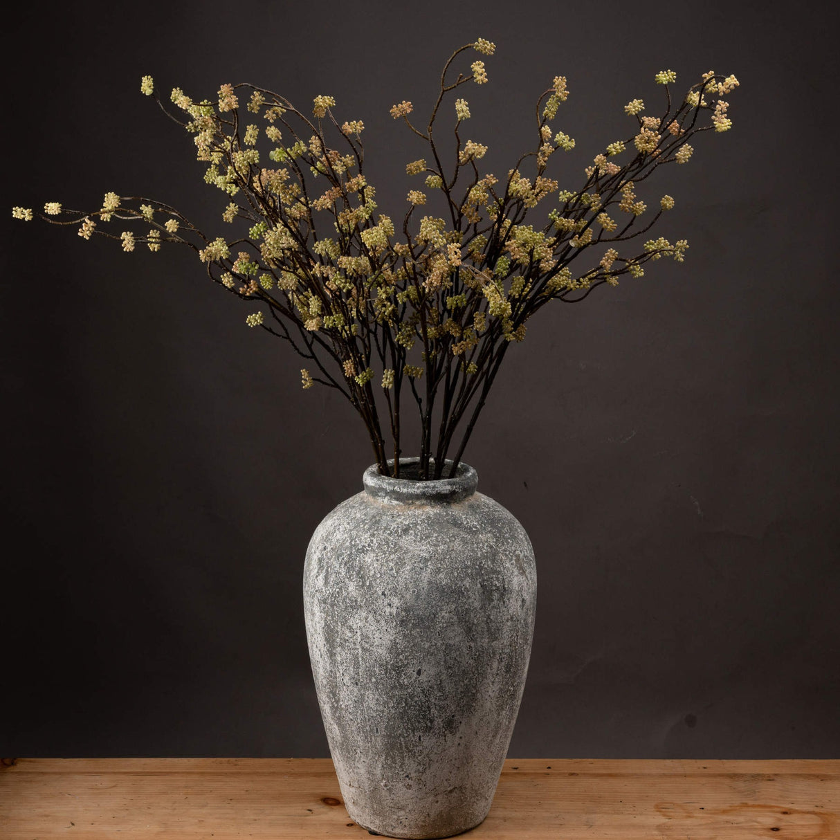 Hill Interiors Branch in Bloom Filler faux stems with clustered green and cream blooms arranged in a weathered grey stone vase against a dark backdrop.