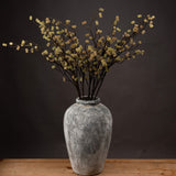 Hill Interiors Branch in Bloom Filler faux stems with clustered green and cream blooms arranged in a weathered grey stone vase against a dark backdrop.