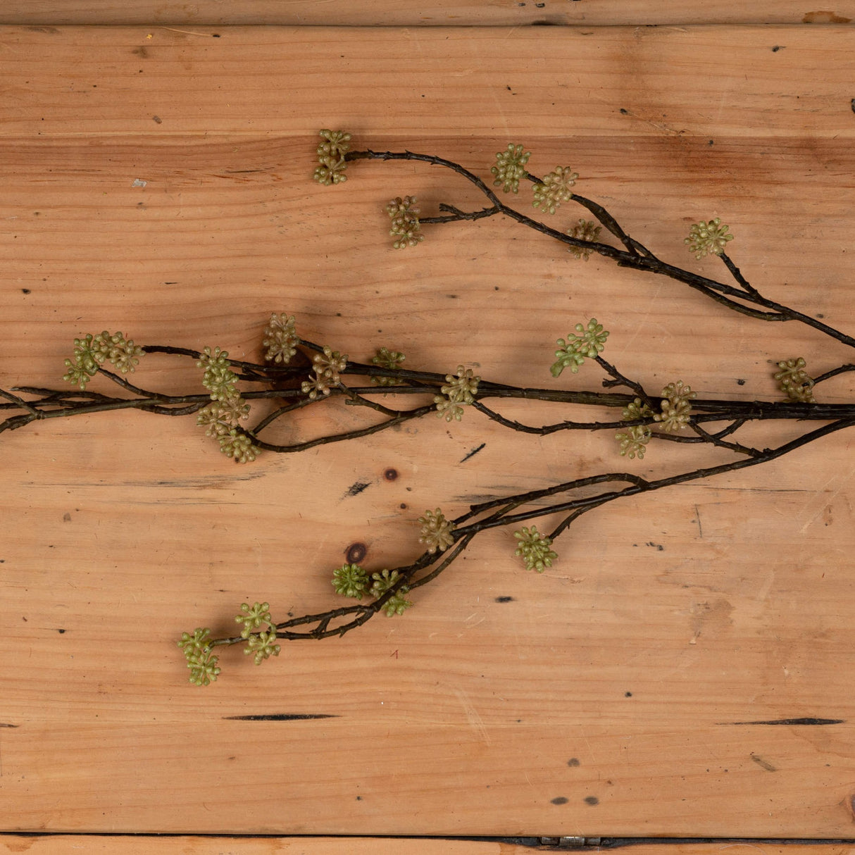 Hill Interiors Branch in Bloom Filler artificial stem with slender dark branches and clusters of small green blooms, laid flat on a light wood surface.