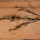 Hill Interiors Branch in Bloom Filler artificial stem with slender dark branches and clusters of small green blooms, laid flat on a light wood surface.