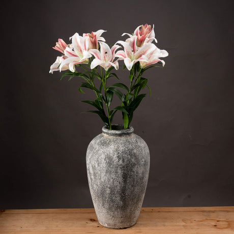 Hill Interiors Stargazer Lily faux stems with white and pink ruffled petals and green foliage, arranged in a weathered stone urn against a dark backdrop.