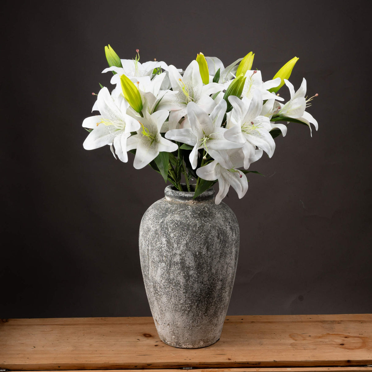 Hill Interiors White Lily faux flower stems with open blooms, green buds, and veined leaves arranged in a weathered grey stone vase on wood.
