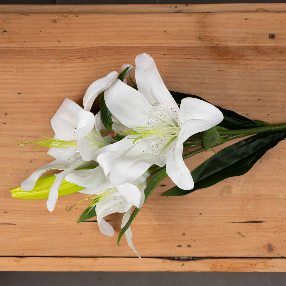 Hill Interiors White Lily faux flower stem with open white blooms, buds, and green veined leaves, displayed on a light wooden surface.