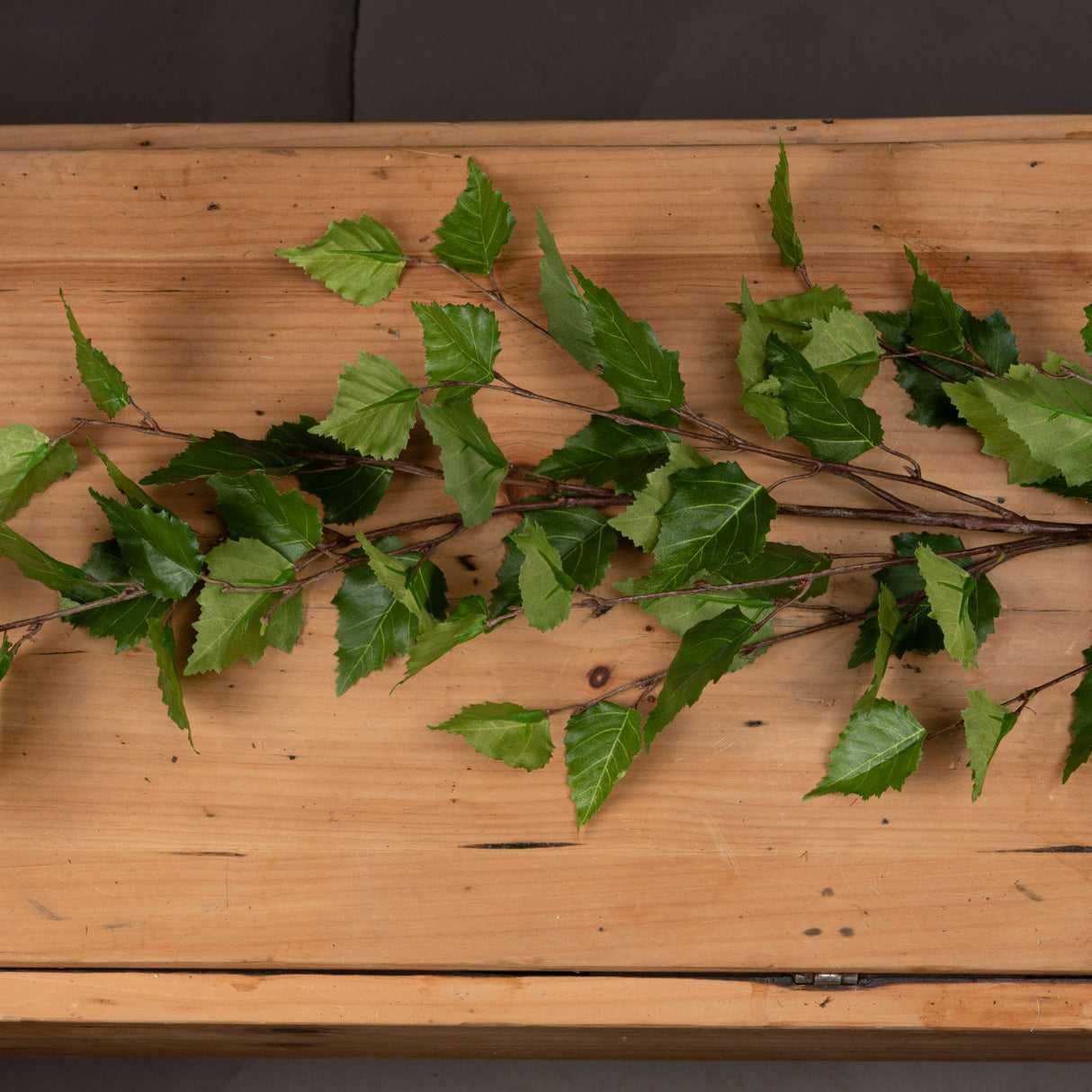 Hill Interiors Green Branch Spray faux foliage stem with slender green leaves fanning along a plastic branch, displayed flat on a rustic wooden surface.