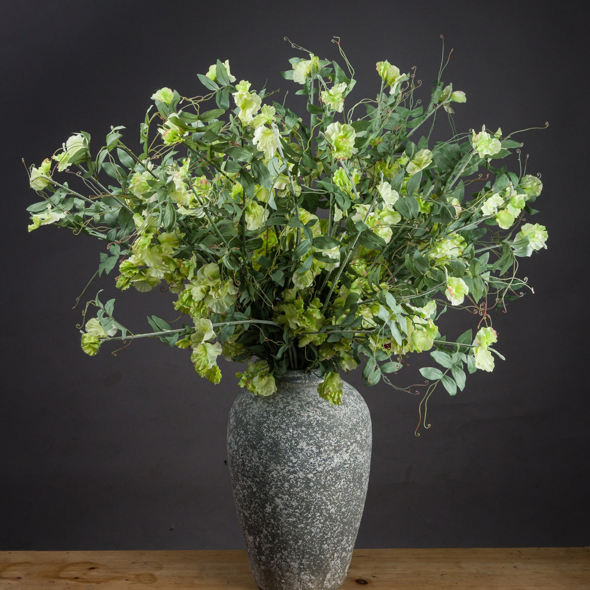 Hill Interiors Sweet Pea Trail faux stems with ruffled green blooms and trailing foliage arranged in a weathered grey stone vase against a dark background.