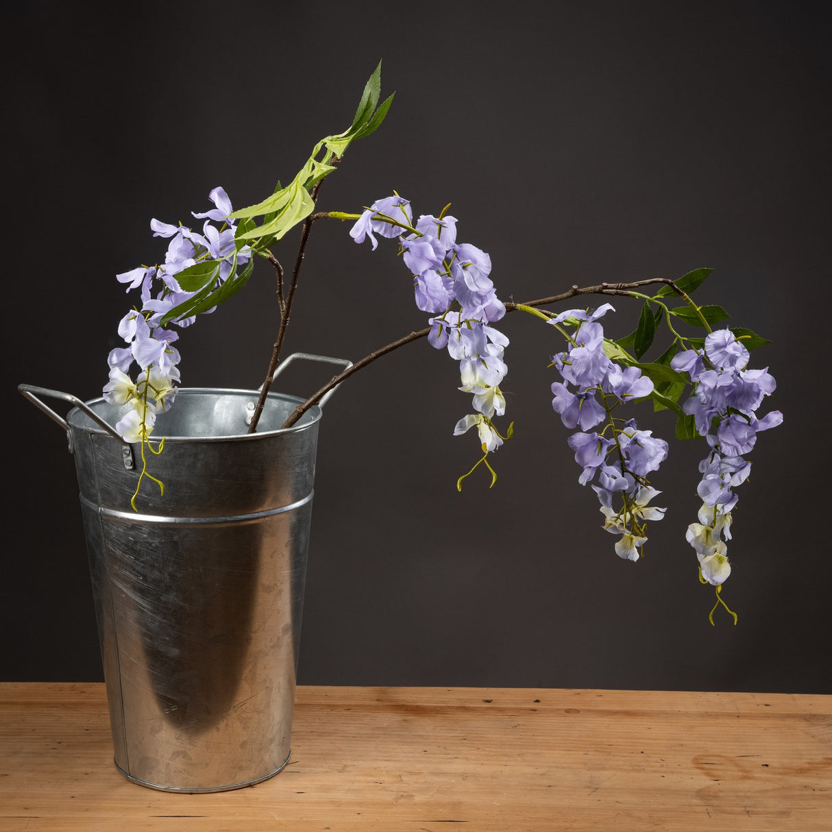 Hill Interiors Lilac Wisteria faux flower stem with cascading pale purple blooms and green leaves, arranged in a galvanised metal bucket on wood.