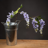 Hill Interiors Lilac Wisteria faux flower stem with cascading pale purple blooms and green leaves, arranged in a galvanised metal bucket on wood.