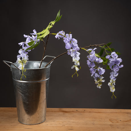 Hill Interiors Lilac Wisteria faux flower stem with cascading pale purple blooms and green leaves, arranged in a galvanised metal bucket on wood.