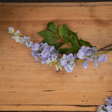 Hill Interiors Lilac Wisteria faux flower stem with cascading pale purple blooms and green leaves, displayed on a light wooden surface.